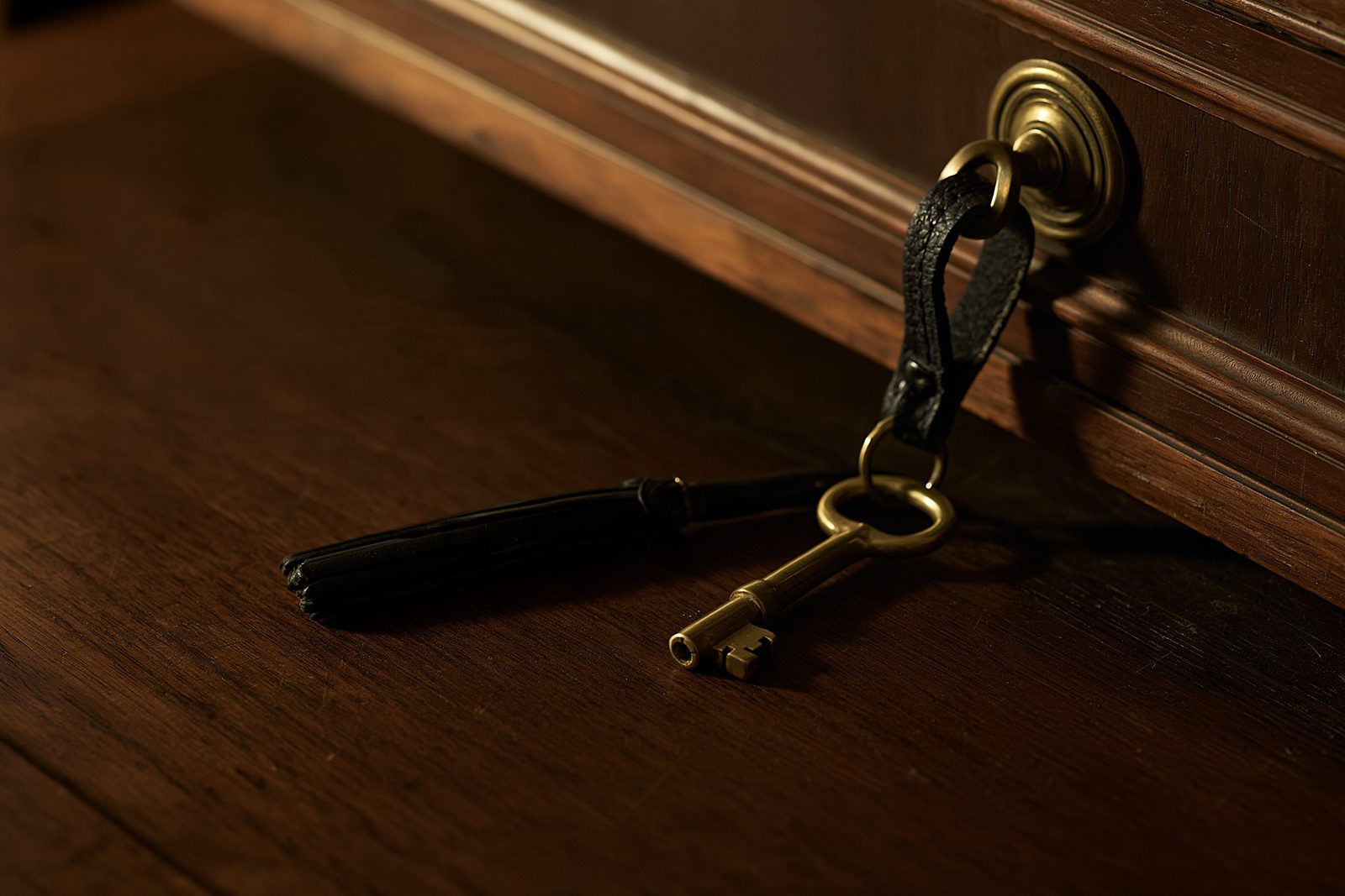 A brass key on a black leather fob hanging from a walnut drawer pull onto the desk, in low warm light.