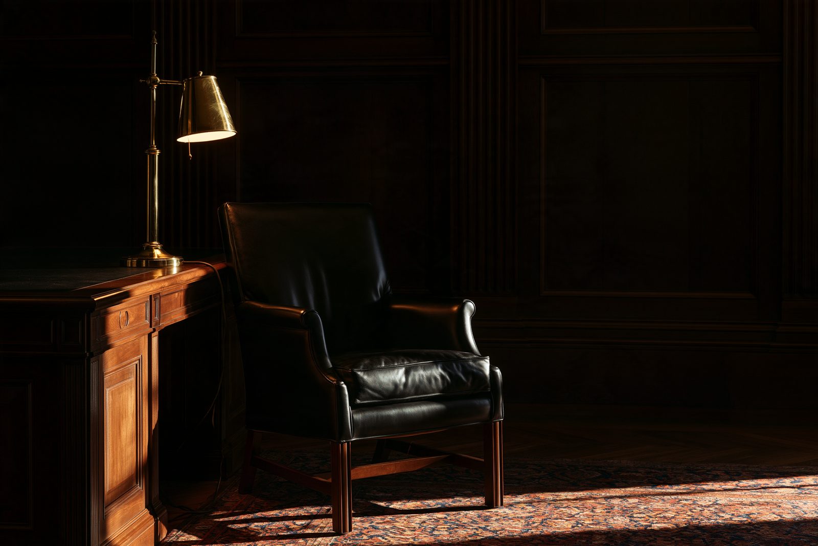 A brass desk lamp beside an empty black leather chair in a dark wood-panelled office, in low warm light.